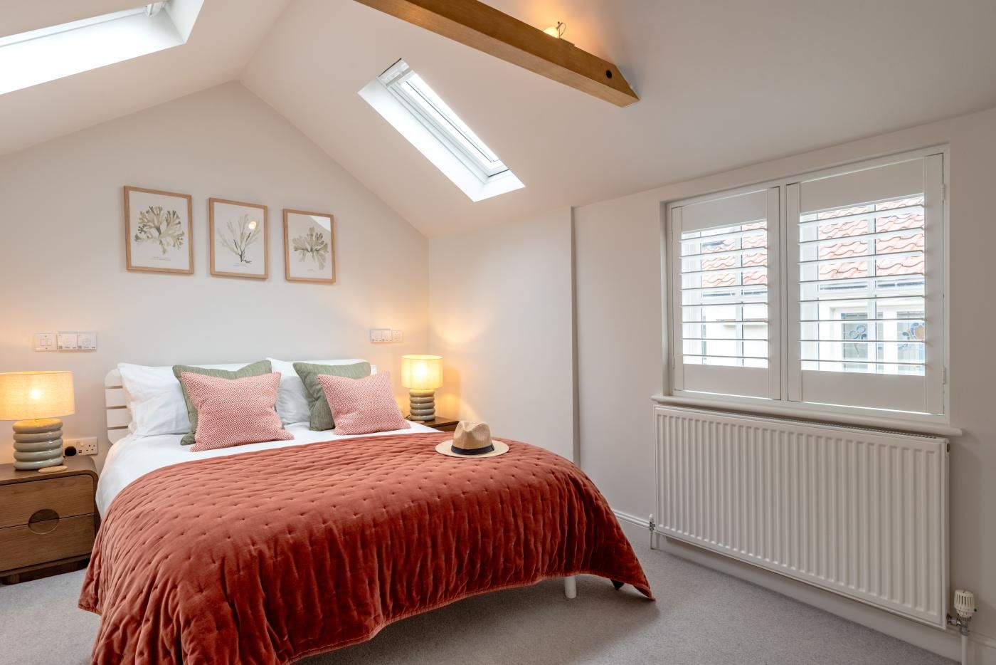 A bedroom with a sloped ceiling, skylight, a bed with a red cover, two bedside lamps, and a window with blinds.