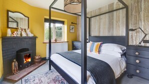 Ein Bett mit Baldachin und schwarzem Gestell, ein Kamin mit sichtbarem Feuer, ein Fenster mit Blick ins Grüne und eine Wandverkleidung aus Holz.
