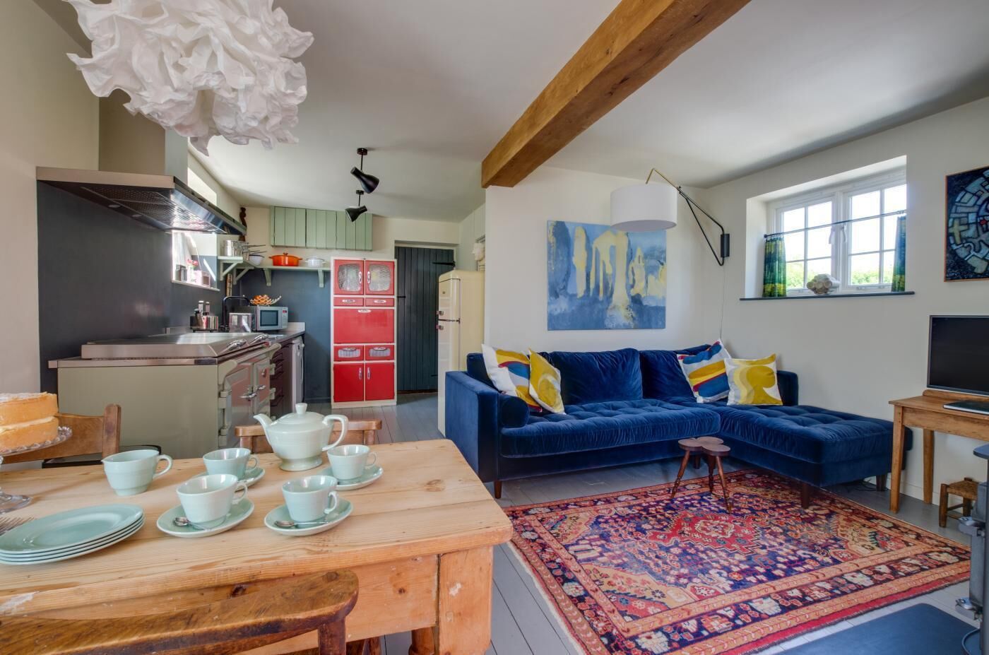 A living room with a blue sofa, a dining table with plates and cups, a kitchen with a stove and cabinets, and a large abstract painting on the wall.
