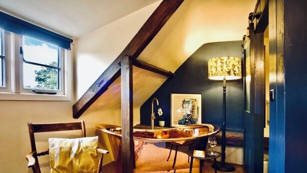 Ein gemütliches Zimmer mit schrägen Wänden, einem Holzstuhl, einem kleinen Tisch, einer Lampe und einem Fenster.
