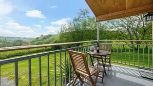 A balcony with a wooden table and chairs, overlooking a green landscape with hills and trees.