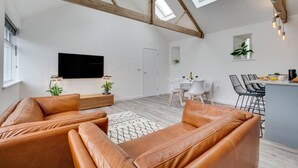 A modern living room with a high ceiling, exposed wooden beams, a flat-screen TV, leather sofas, a dining area with a white table and chairs, and a kitchen with a bar counter. - 1 Bed in Mitchell (oc-p00750) (Mitchell)