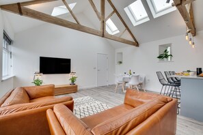 A modern living room with a high ceiling, exposed wooden beams, a flat-screen TV, leather sofas, a dining area with a white table and chairs, and a kitchen with a bar counter. - 1 Bed in Mitchell (oc-p00750) (Mitchell)