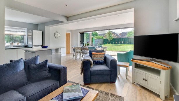Ein modernes Wohnzimmer mit Sofa, Sessel und Couchtisch. Es gibt einen Essbereich mit Tisch und Stühlen sowie eine Küche mit Kühlschrank und Spüle. Ein Fernseher ist an der Wand montiert und ein großes Fenster bietet einen Ausblick ins Freie.