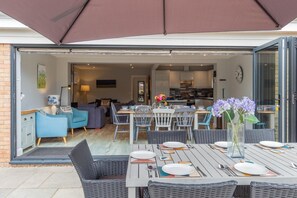 An open-plan living area with a dining table set for four, a kitchen in the background, and a large umbrella providing shade.
