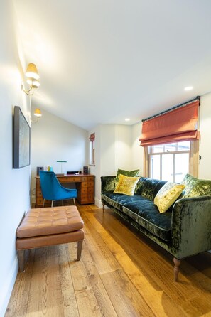 A living room with a dark green sofa, a blue chair, a wooden coffee table, a desk with a computer, and a window with a red blind. - 1 Bed in Southwold (oc-med) (Southwold)