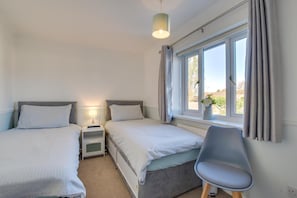 Ett sovrum med två sängar, ett skrivbord, en stol, ett nattduksbord med en lampa och ett fönster med gardiner.