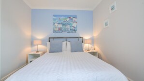 A bedroom with a large bed, two bedside tables with lamps, a painting on the wall, and a small shelf.