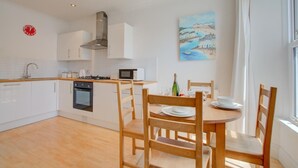 A modern kitchen with white cabinets, a gas stove, and a microwave. There is a dining table with chairs and a painting of a coastal scene on the wall.