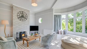 A modern living room with a fireplace, a large window with curtains, a round seating area, and a television on a stand. - 3 Bed in Trevone (oc-p00758) (Trevone)
