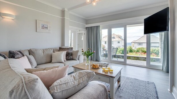 A spacious living room with a large sofa, a coffee table with breakfast items, and a view of the outside through large windows. - 3 Bed in Trevone (oc-p00758) (Trevone)