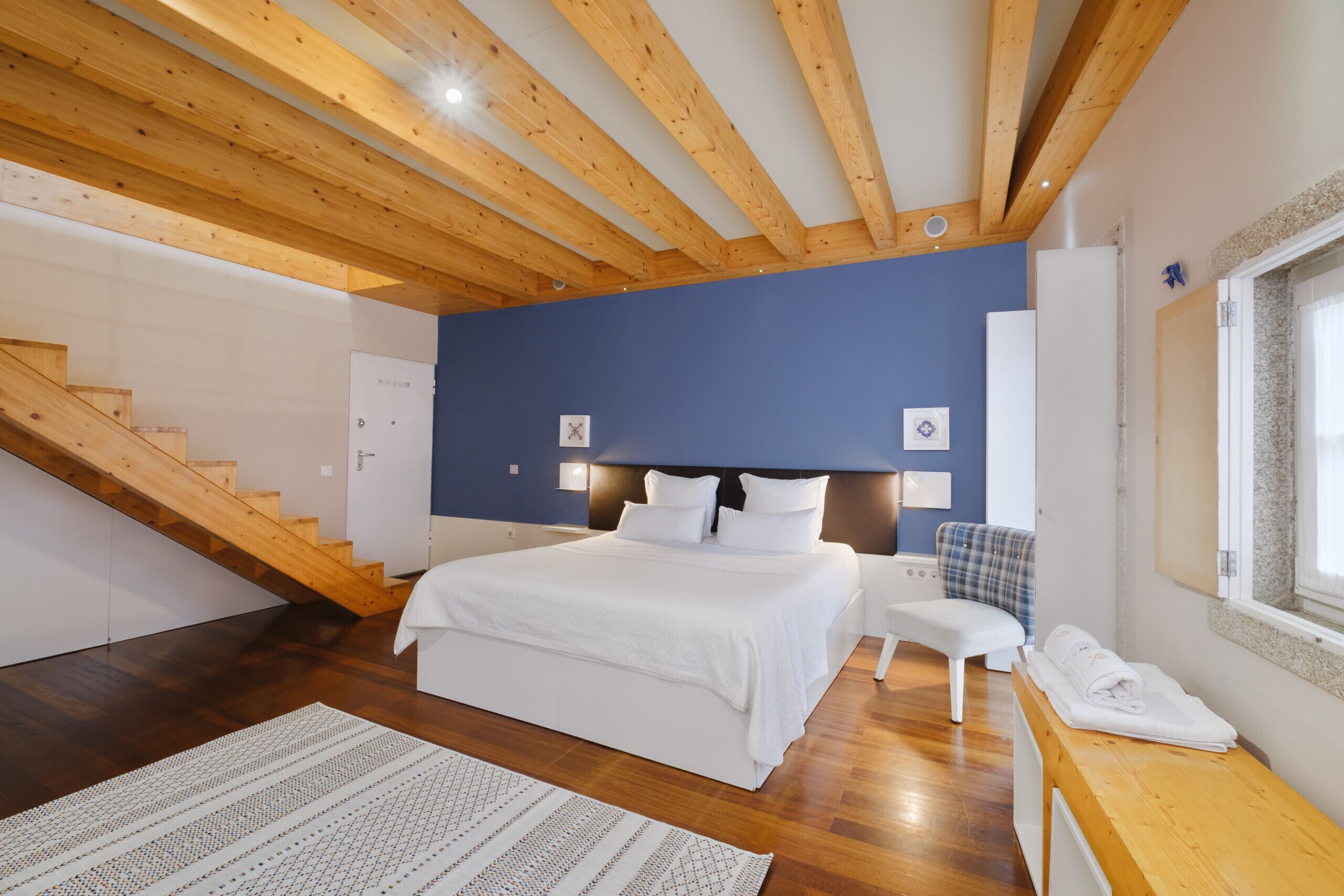 A bedroom with a wooden ceiling, a bed with white bedding, a chair, a desk, and a window.