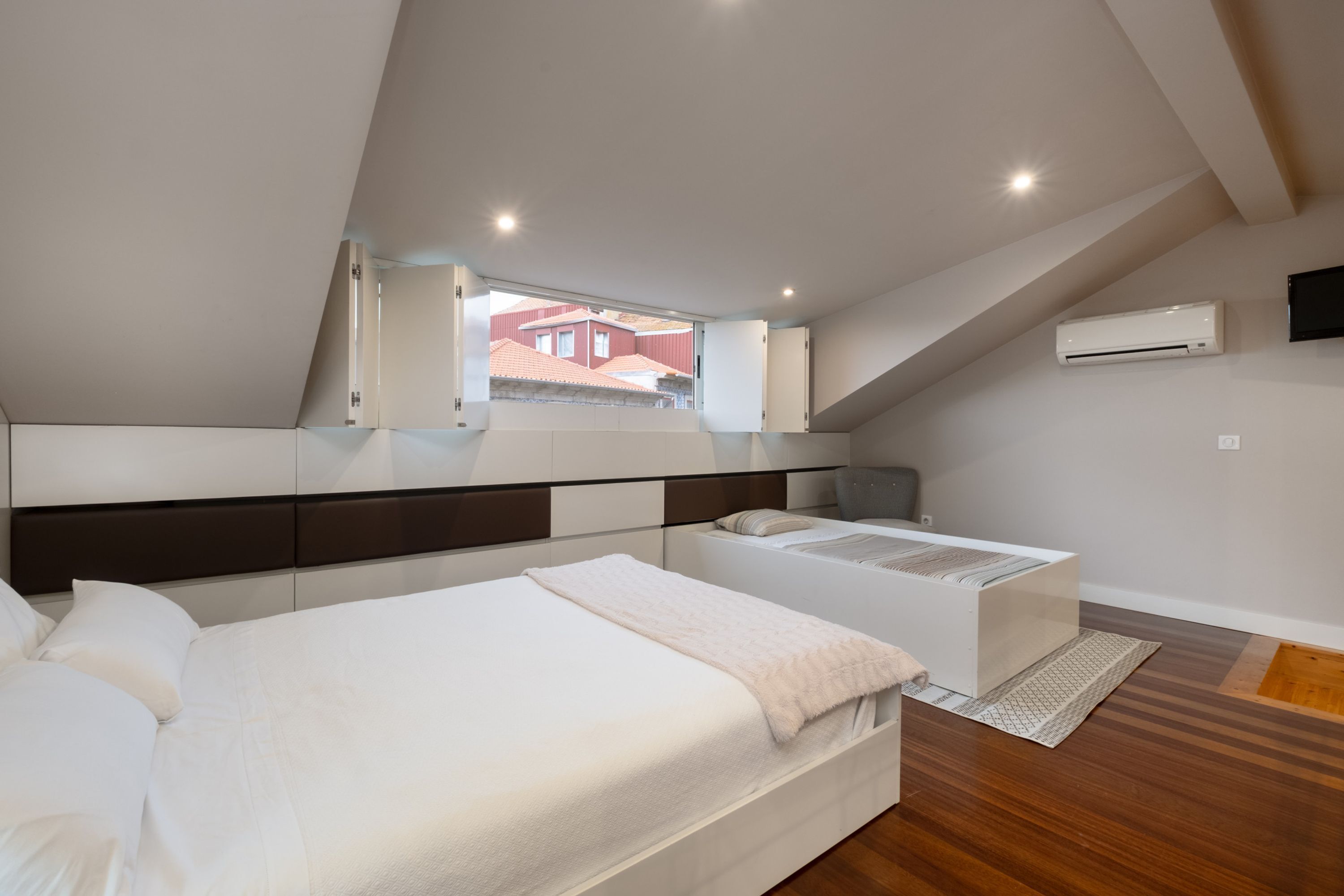 A bedroom with a bed, a bedside table, a chair, a window, and an air conditioning unit.