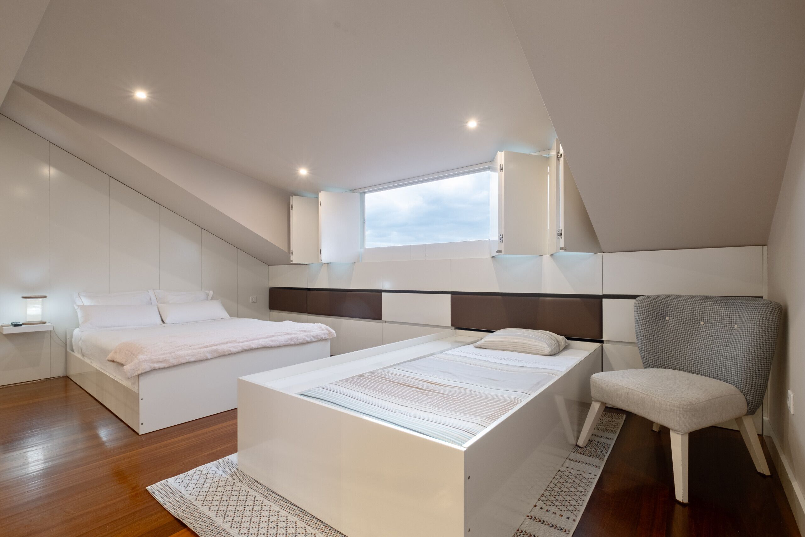 A modern bedroom with a large bed, a smaller bed, a chair, and a window.