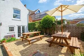 A patio area with wooden picnic tables and a large umbrella, surrounded by a gravel garden and a white building. - 6 Bed in Cromer (oc-1996) (Cromer)