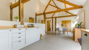 Una cocina amplia con armarios blancos, vigas de madera, un comedor con mesa y sillas, y vista a otra habitación.