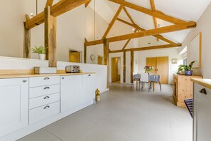 Une cuisine spacieuse avec des meubles blancs, des poutres en bois, un coin repas avec une table et des chaises, et une vue sur une autre pièce.