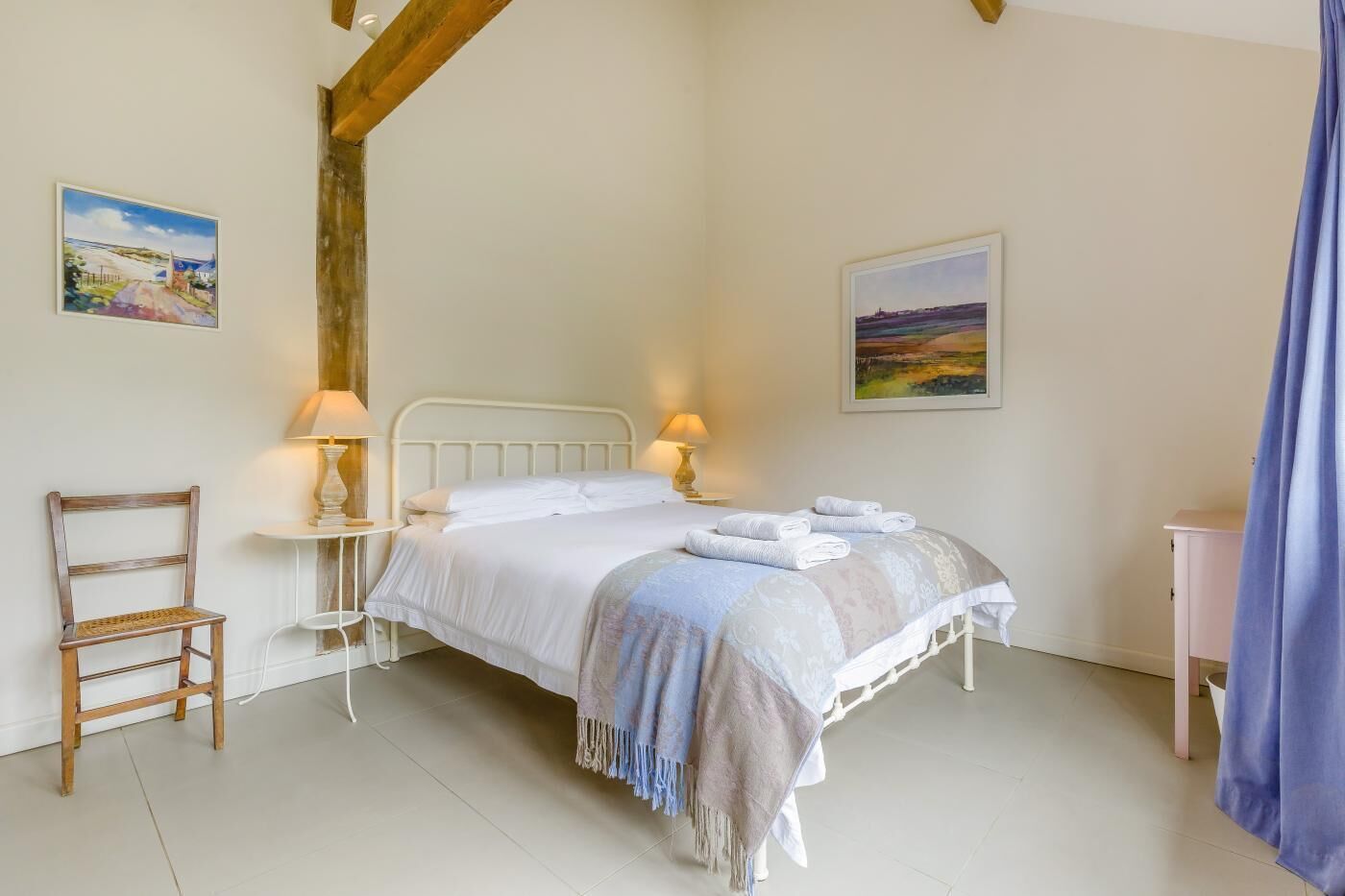 A bedroom with a bed, two bedside lamps, a chair, and two paintings on the wall.