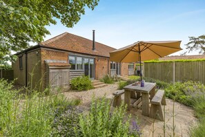 Une maison en briques avec un toit en tuiles, une cheminée, une clôture en bois et un jardin avec une table et des chaises placées sous un grand parasol.