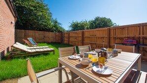 A backyard with a wooden table set for breakfast, a grill, and a fenced area with lounge chairs. - 2 Bed in Mundesley (oc-1991) (Mundesley)