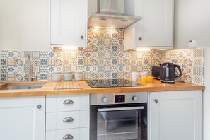 A kitchen with a tiled backsplash, a stainless steel range hood, a digital clock, and a kettle on the counter. - 1 Bed in Abergele (oc-bow134) (Llanfair Talhaiarn)