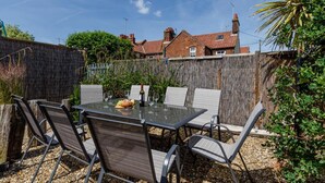 An outdoor patio with a glass table and metal chairs, surrounded by a wooden fence and greenery. - 4 Bed in Mundesley (oc-1997) (Mundesley)