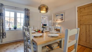 A dining area with a wooden table set for four, surrounded by chairs, a pendant light, and a view through a window. - 4 Bed in Mundesley (oc-1997) (Mundesley)