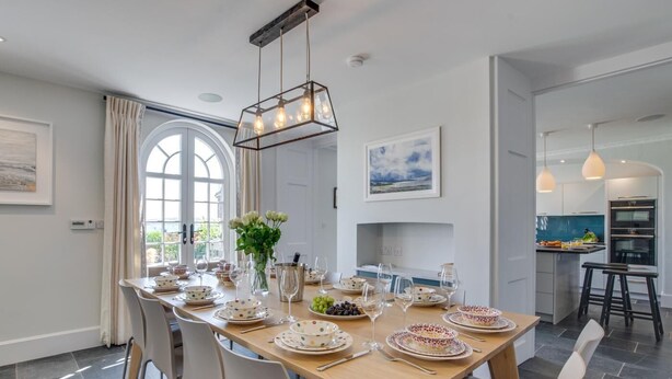 Un comedor con una mesa larga dispuesta para una comida, una cocina al fondo y vistas a través de un gran ventanal.