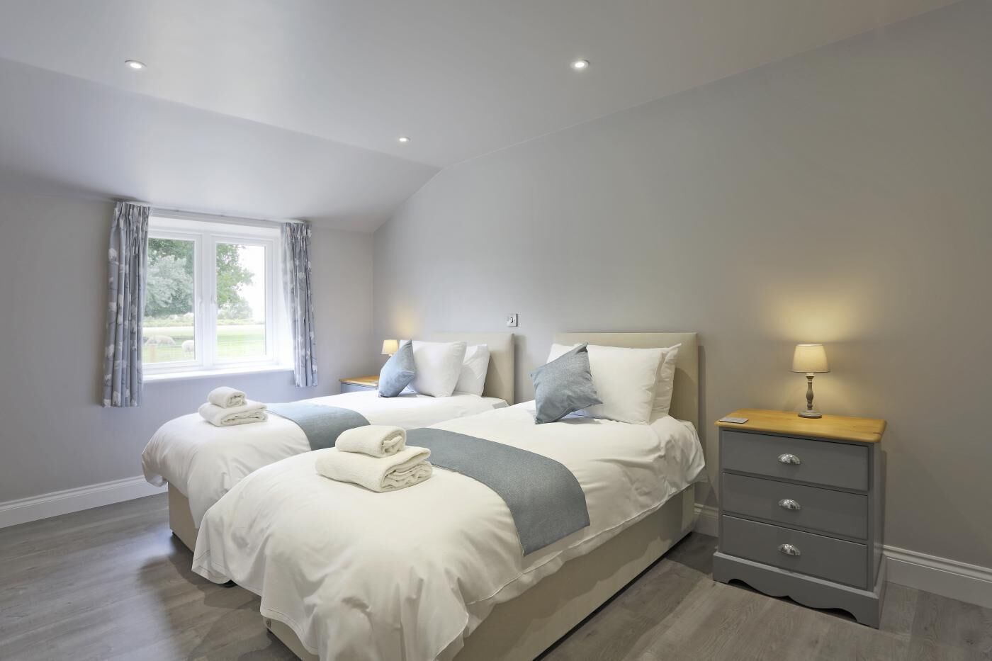 Een moderne slaapkamer met twee bedden, een nachtkastje met een lamp, en uitzicht op het groen door een raam.