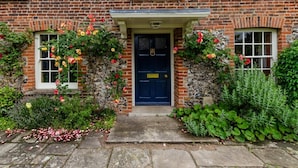 Ein Backsteinhaus mit einer dunkelblauen Tür, einer Steinmauer und einem kleinen Garten mit Blumen und Sträuchern.
