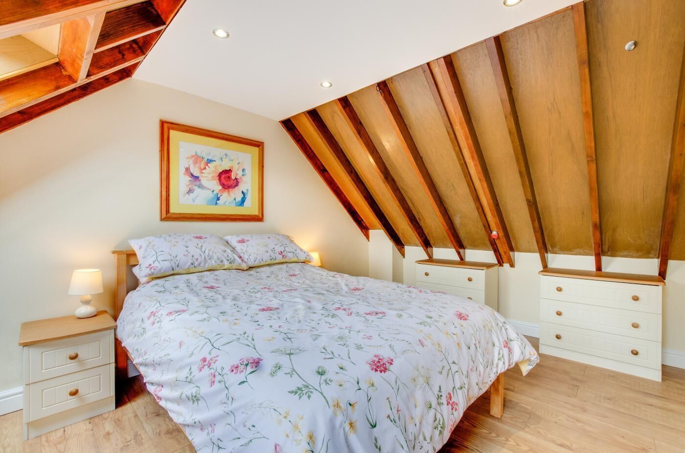 Une chambre avec un plafond en pente en bois, un lit avec une literie à motifs floraux, une table de chevet et une commode.