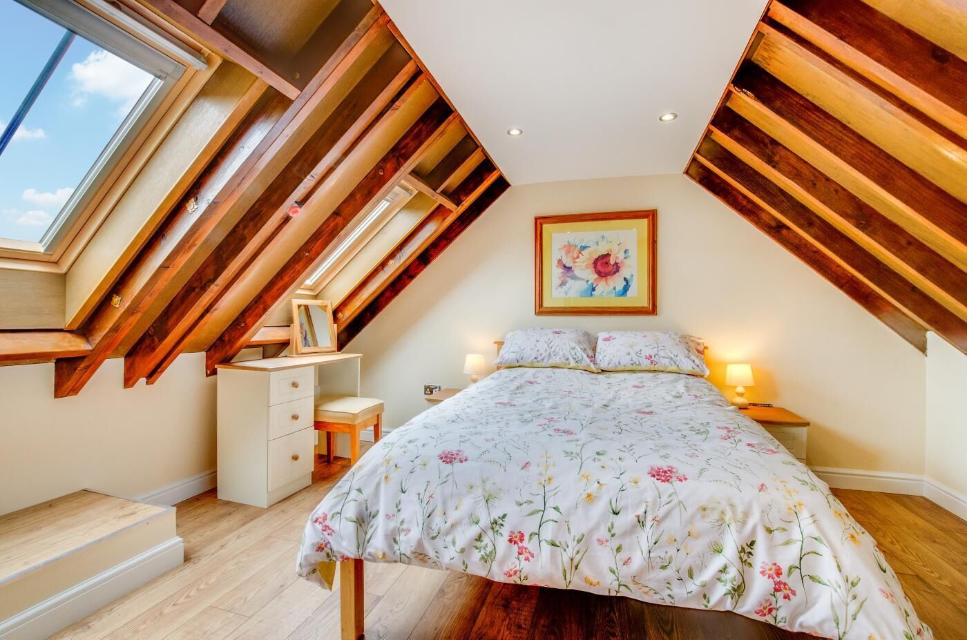 A bedroom with a sloped ceiling, wooden beams, a bed with floral bedding, a bedside table with a lamp, and a framed picture on the wall.