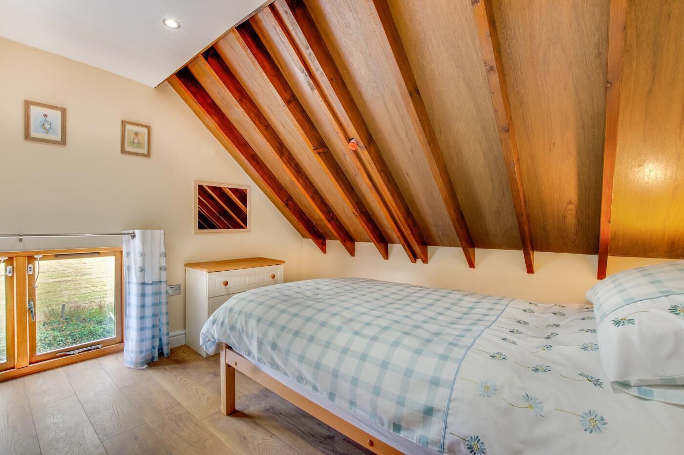 A bedroom with a sloped ceiling, a bed with a checkered bedspread, a bedside table, and a window with curtains.