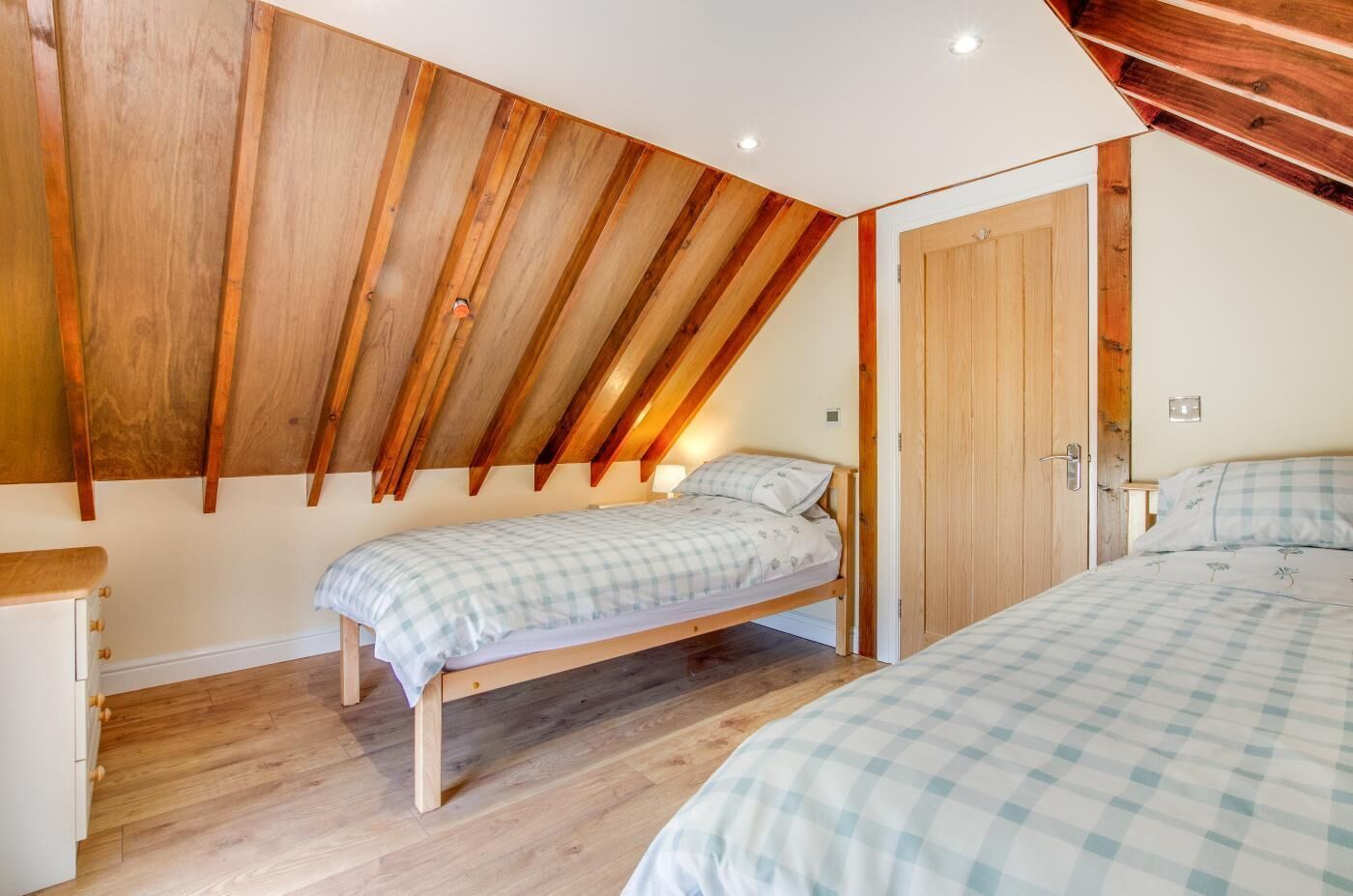 A bedroom with a sloped ceiling, wooden beams, two beds with checkered bedding, a wooden dresser, and a door.