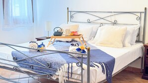 Una cama bien hecha con estructura metálica, sábanas blancas y una manta azul y blanca a rayas. Sobre la cama, un cesto de madera con una tetera, taza y un pequeño ramo de flores.