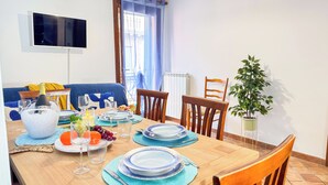 Un comedor con una mesa de madera dispuesta para una comida, rodeada de sillas, un televisor montado en la pared y una planta en maceta.