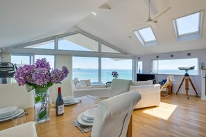 Un amplio salón con un ventanal grande con vistas al mar, una mesa de comedor dispuesta con platos y una botella de vino, y un telescopio sobre un trípode.