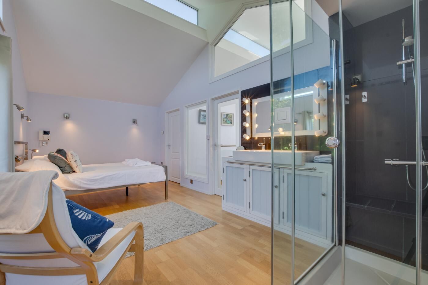 Ein modernes Schlafzimmer mit einem großen Bett, einer Sitzecke, einer gläsernen Dusche und einer Küchenzeile mit Waschtisch und Beleuchtung.