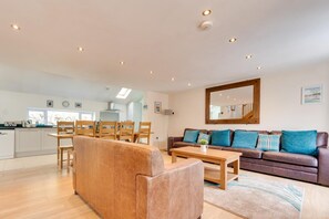 Uma sala de estar espaçosa com um sofá de couro marrom, uma mesa de centro de madeira e um sofá de couro marrom com almofadas verde-água. É visível uma cozinha ao fundo, com armários brancos e uma janela.