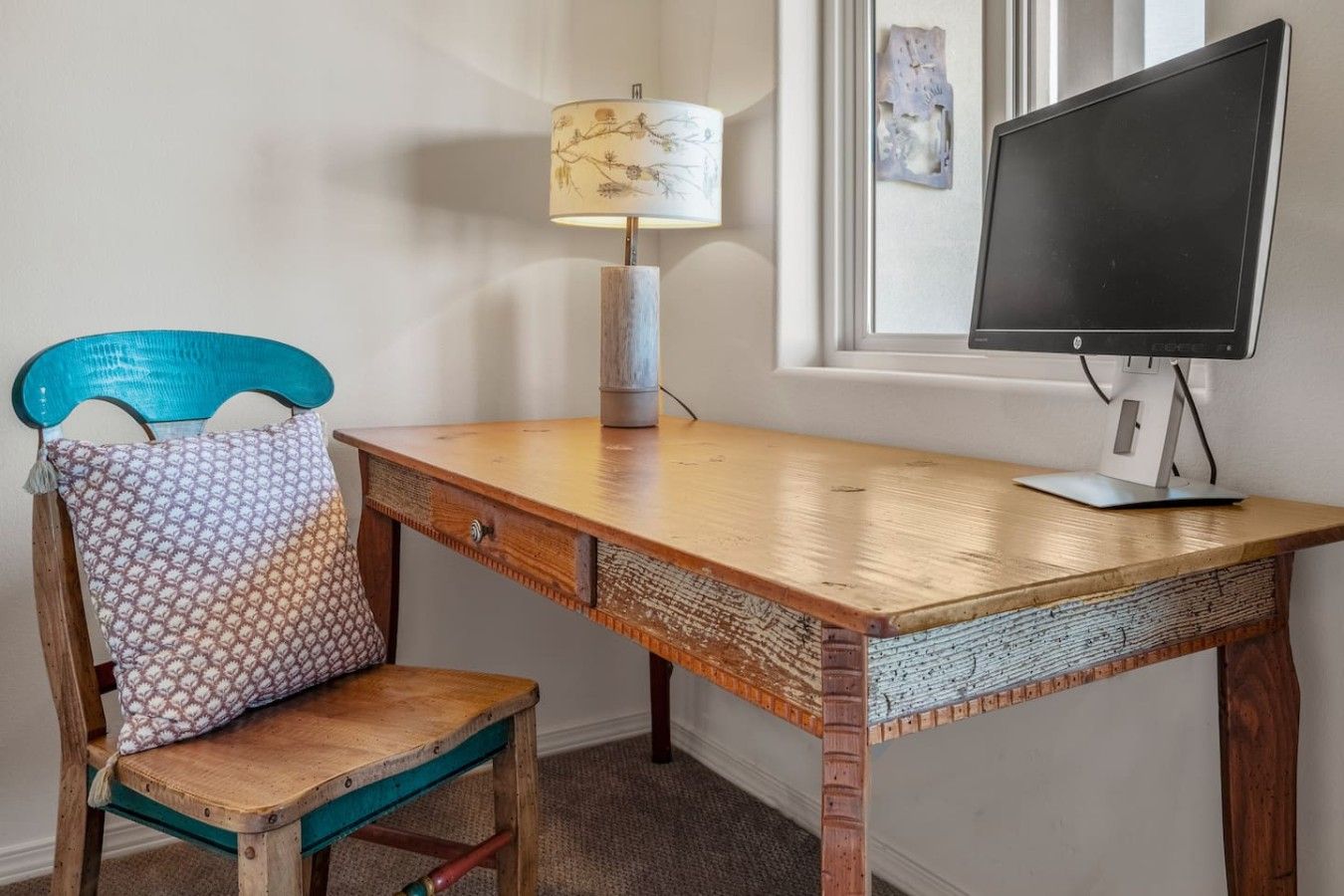Uma mesa de madeira com um monitor, uma lâmpada e uma cadeira com almofada estampada.