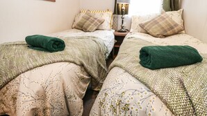 A twin room with two beds, each with a green towel, floral-patterned bedding, a nightstand with a lamp, and a window with curtains. - Loch Lomond Holiday Home (Arrochar)