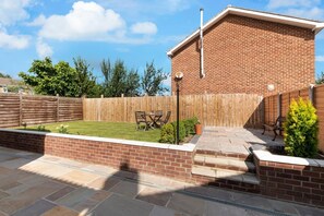 A backyard with a brick house, a wooden fence, a patio with a table and chairs, and a small garden area. - 4 Bed in Whitby (oc-w466) (Whitby)