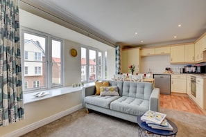 A modern apartment with a grey sofa, a dining table, and a kitchen area. - 2 Bed in Whitby (oc-w463) (Whitby)