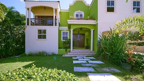 A two-story house with a green and white facade, a balcony on the upper floor, and a well-maintained garden with stepping stones leading to the entrance. - Serenity Villa - by Zenbreak (Speightstown)