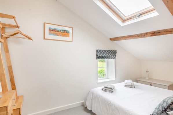 Ein Schlafzimmer mit Dachfenster, einem Bett mit ordentlich gefalteten Handtüchern, einem Holzstuhl, einem kleinen Fenster mit Vorhang und einem gerahmten Bild an der Wand.