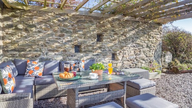 Un muro de piedra con una pérgola de madera, un sofá de mimbre con cojines, una mesa de vidrio con alimentos para el desayuno y una planta en maceta.