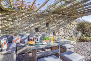 Un muro de piedra con una pérgola de madera, un sofá de mimbre con cojines, una mesa de vidrio con alimentos para el desayuno y una planta en maceta.