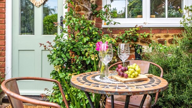A round outdoor table with a mosaic design, surrounded by greenery and a brick wall. - 2 Bed in Heacham (oc-2013) (Heacham)