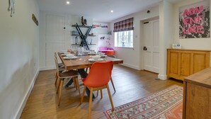 A dining area with a wooden table set for four, surrounded by wooden chairs. There is a window with a plaid curtain, a radiator, and a shelf with decorative items. - 4 Bed in Walberswick (oc-4ms) (Walberswick)