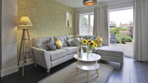 Ein Wohnzimmer mit grauem Sofa, einer Stehlampe, einem runden Couchtisch mit einer Vase voller Sonnenblumen und Blick ins Freie durch große Fenster.